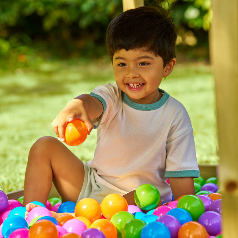 Jeune enfant assis dans une piscine à balles colorées en extérieur, souriant et tenant une balle orange.