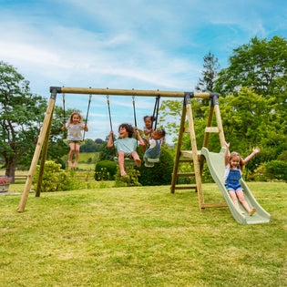 Cinq enfants jouent sur un portique en bois dans un jardin. Quatre d’entre eux se balancent sur les balançoires, tandis qu’un autre profite de la structure avec échelle et toboggan. La scène se déroule sur une pelouse verdoyante, entourée d’arbres, sous un ciel bleu.