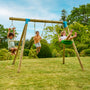Trois enfants jouent sur un portique en bois dans un jardin. L’un utilise une échelle corde, tandis que deux autres se balancent sur une balançoire. Le décor montre une pelouse verte entourée d’arbres et d’arbustes, sous un ciel bleu.