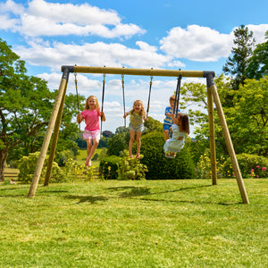 Quatre enfants jouent sur un portique en bois installé dans un jardin. Deux enfants se balancent chacun sur une balançoire simple, tandis que deux autres sont sur un vis-à-vis. La scène se déroule sur une pelouse ensoleillée entourée d’arbres et d’arbustes, sous un ciel bleu parsemé de nuages.