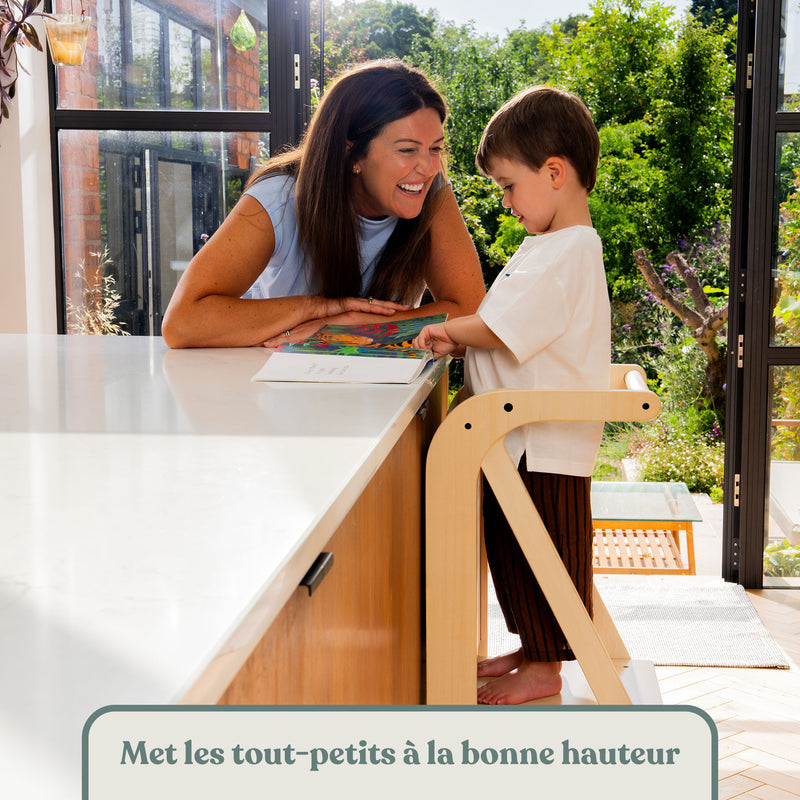 Enfant sur la tour d'apprentissage Montessori en bois, lisant un livre avec sa maman au plan de travail