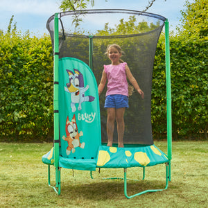 Enfant sautant dans un trampoline décorée de personnages Bluey avec filet de sécurité noir, installé dans un jardin avec pelouse et haies en arrière-plan.