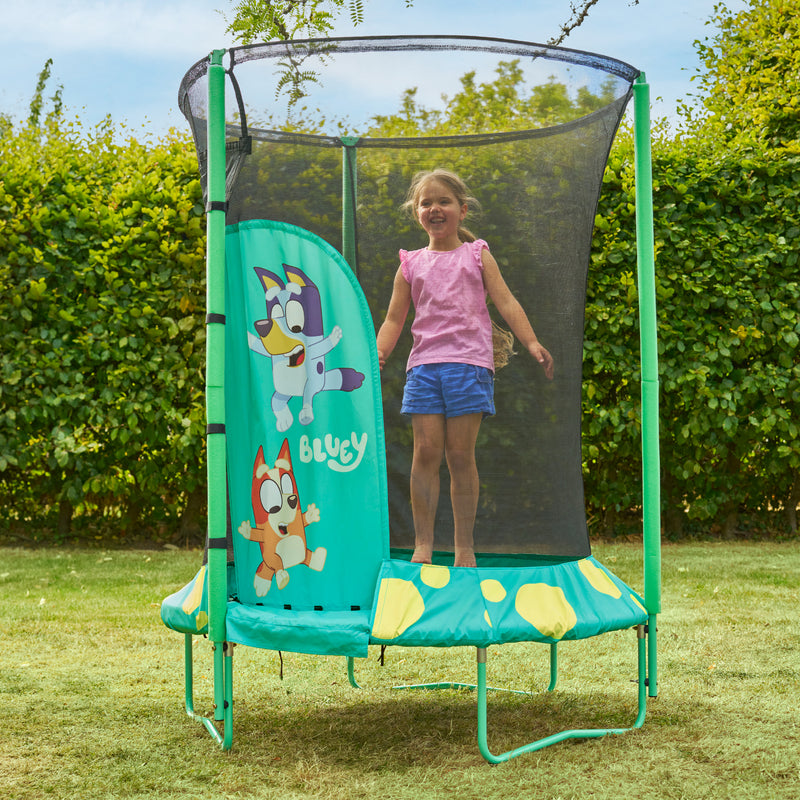 Enfant sautant dans un trampoline décorée de personnages Bluey avec filet de sécurité noir, installé dans un jardin avec pelouse et haies en arrière-plan.