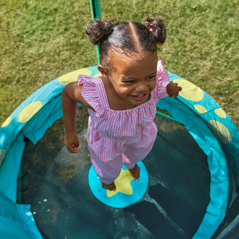Vue plongeante d’un enfant sautant à l’intérieur d’un trampoline, avec tapis de saut noir et protection rembourrée turquoise à motifs jaunes, installé sur une pelouse.