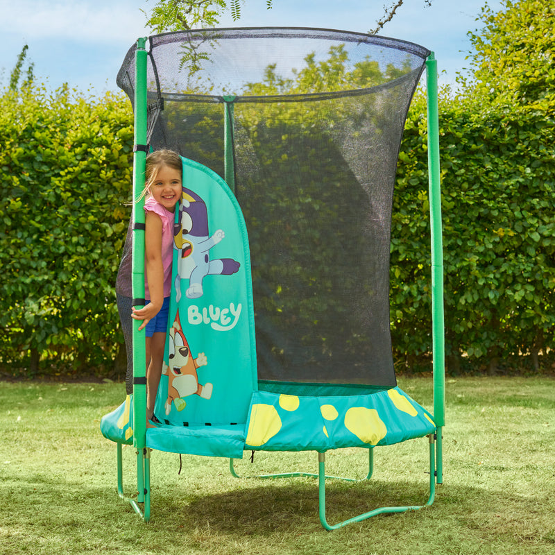 Enfant se tenant à l’entrée d’un trampoline décorée de personnages Bluey, avec filet de sécurité noir, installé dans un jardin avec pelouse et haies en arrière-plan.