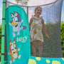 Détail d’un trampoline pour enfant avec filet de sécurité noir et protection rembourrée turquoise décorée de personnages Bluey, avec un enfant debout à l’intérieur, installé dans un jardin.