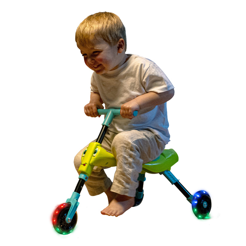 Un enfant souriant roule sur un tricycle vert et bleu avec des roues lumineuses.