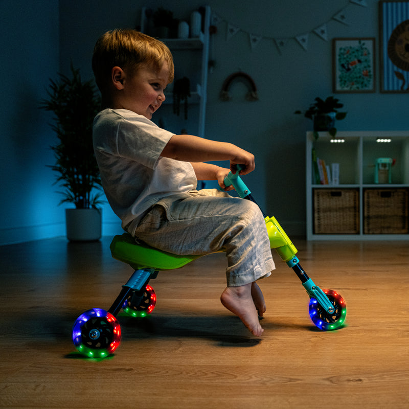Un enfant roule sur un tricycle vert et bleu à l’intérieur, avec des roues LED multicolores allumées.